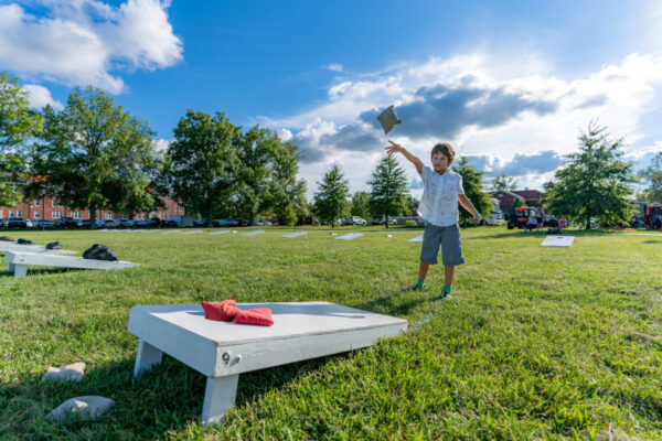 A young boy is tossing bean bags at a corn hole board during a summer afternoon with some clouds in the sky.