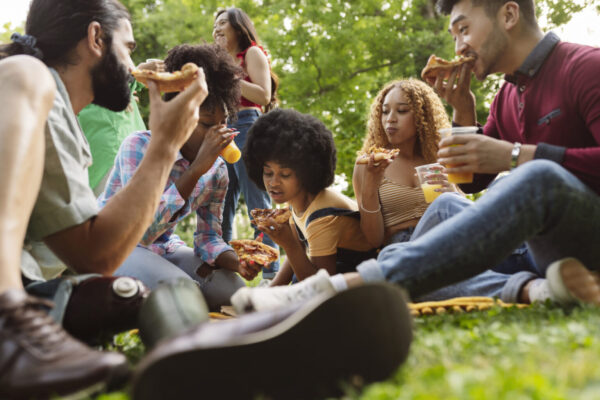 Group of closed friends enjoying dinner in park. Man and women sitting on plaid around pizza and bottles of beer, taking slices from box and eating. Takeaway dinner concept