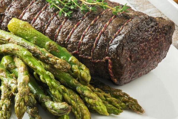 Whole beef tenderloin cooked in the oven, tied with a rope in a white porcelain plate, and the asparagus next to it.