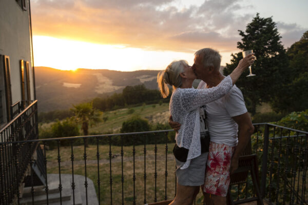 A senior couple standing outside the villa they are staying at in Tuscany, Italy. They are kissing with the landscape and sunset behind them. The woman is holding a wine glass.