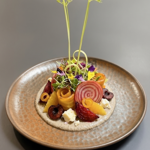 Plated gourmet salad featuring roasted red, golden, and striped chioggia beets, fresh orange segments, dark pickled cherries, and Brillat-Savarin cheese arranged over a creamy puree, topped with microgreens and two tall herb shoots on a textured bronze ceramic plate.