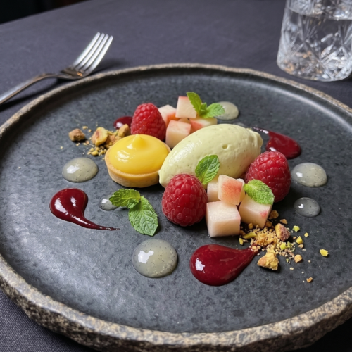 Deconstructed gourmet dessert featuring a mini lemon tartlet, a quenelle of light green pistachio cream, fresh red raspberries, diced white peach, mint leaves, and dots of vanilla gel and berry sauce on a dark slate plate.