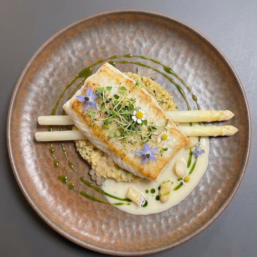 Pan-seared flounder fillet resting on a bed of cornbread dressing, with white asparagus spears placed horizontally behind the fish, finished with white asparagus caviar, green basil oil drizzle, trinity beurre blanc sauce, and delicate microgreens on a bronze ceramic plate.