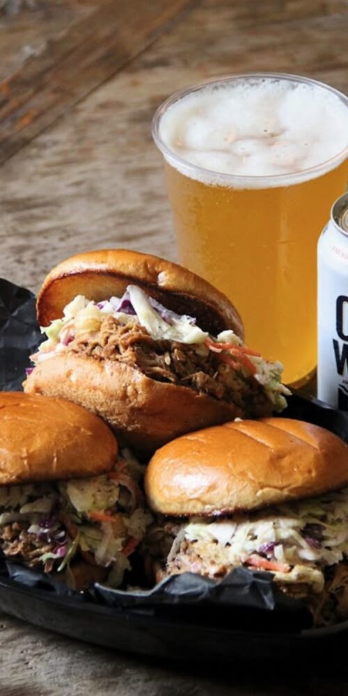 Three BBQ pulled pork sandwiches topped with coleslaw on toasted brioche buns, served next to a City Wide pale ale beer.